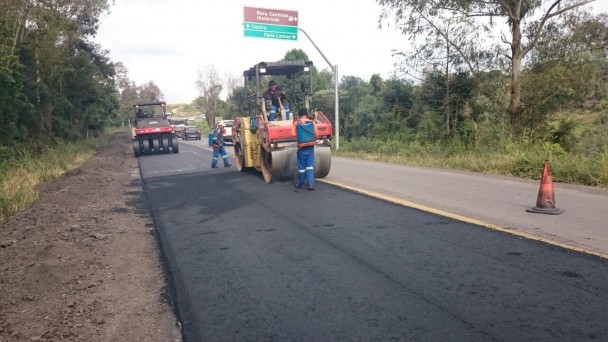 A foto mostra em trecho da BR-470 sendo recapeado em metade da pista. Um rolo compressor passa pela via, dirigido por um operário, acompanhado de outros dois a pé. Uma placa turística aparece mais ao fundo, assim como outra máquina de obras.