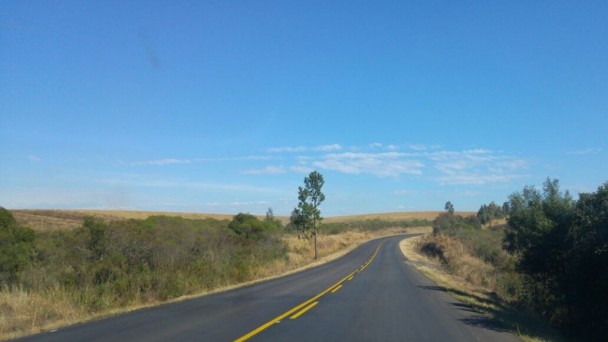 A foto mostra um trecho da ERS-122 em reta, com uma curva para a direita mais adiante. O asfalto está recuperado, em boas condições e com a faixa pintada no meio da pista. Às margens, vegetação com mato e árvores e céu azul com poucas nuvens.