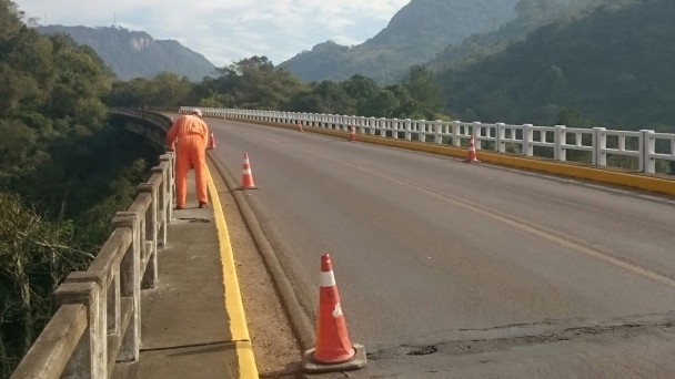 Técnico do Daer, uniformizado,realiza pintura no guarda-corpo de ponte, cercada de vegetação e montanhas; há cones de sinalização na pista.