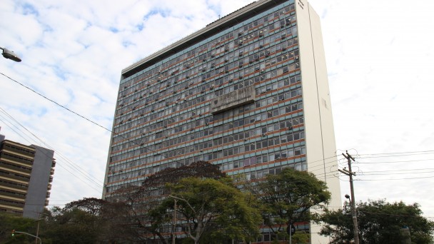 A foto mostra a fachada do prédio do Daer, com árvores e postes de energia elétrica à frente. Ao lado, ao fundo da foto, o Tribunal de Justiça do Estado