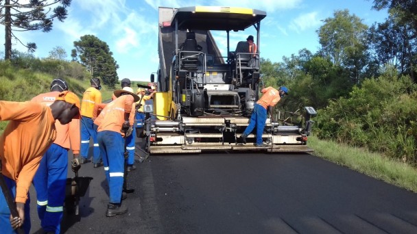 A foto mostra uma equipe de, aproximadamente, sete operários, trabalhando no recapeamento de uma estrada pavimentada. O grupo utiliza carrinhos de mão, pás e uma máquina que ocupa meia pista da via.