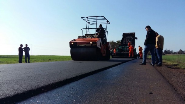 A foto mostra obras de recapeamento na rodovia ERS-223. Na imagem, uma máquina passa pela recém colocada camada de asfalto na pista, com outros operários trabalhando e pessoas olhando.