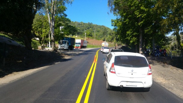 A foto mostra um trecho da BR-470, com tráfego de veículos, onde há pavimento e sinalização restaurados. às margens, árvores e propriedades rurais.