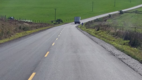 A foto mostra a rodovia ERS-126 com asfalto e pintura renovados. Ao longe, um caminhão faz a curva na estrada, em sentido oposto à câmera. Às margens da estrada, vegetação e cercas de propriedades rurais.