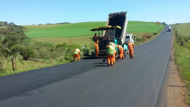 A foto mostra um grupo de operários trabalhando na recuperação do asfalto da ERS-467. Um caminhão descarrega material asfáltico enquanto outra máquina faz a compactação na pista. Ao fundo, caminhões e carros seguem no sentido da frente de obras.