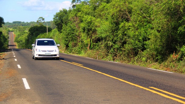 A foto mostra uma grande extensão da ERS-561 já recuperada, com asfalto e sinalização de pista em condição de novos. Um carro branco circula na via. Às margens, vegetação densa.