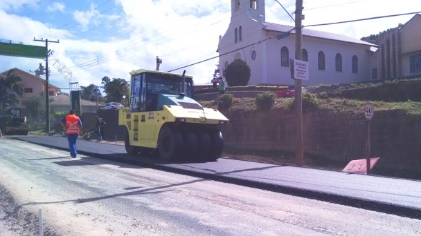 Na foto, um rolo compressor passa sobre a faixa já asfaltada da rodovia ERS-713, na entrada de Sertão Santana. Ao fundo, uma igreja.