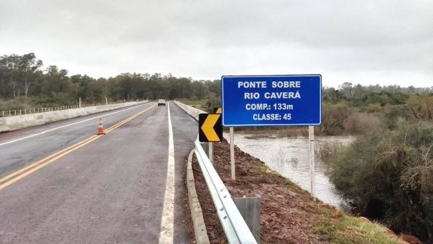 A foto mostra a nova ponte sobre o Rio Caverá, em Alegrete, com destaque para a placa indicativa da estrutura. É possível ver o rio à direita da imagem.