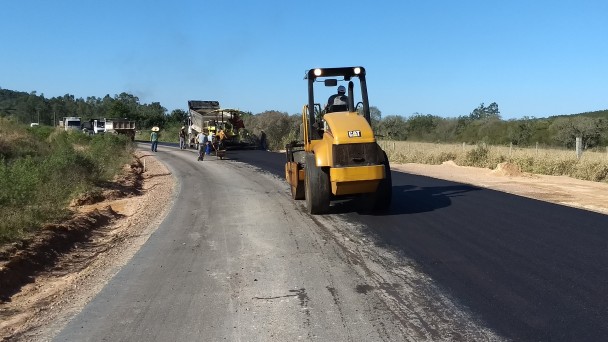 A foto mostra máquinas trabalhando na pavimentação do acesso a Sertão Santana. Uma das vias da estrada está com asfalto recém implantado, enquanto a outra está com as camadas anteriores.