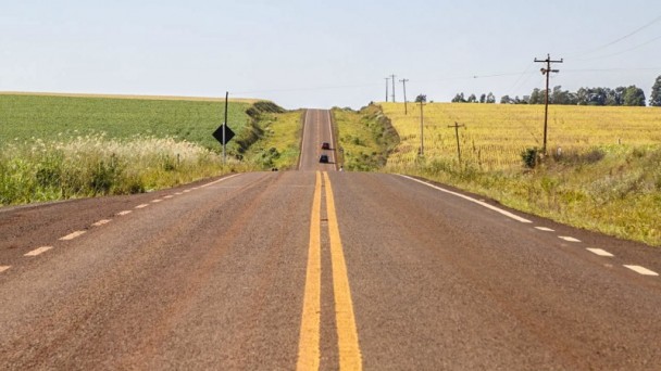 A foto mostra a rodovia de acesso ao município de Eugênio de Castro, já pavimentada e com sinalização pronta. Lavouras e céu azul completam a paisagem.