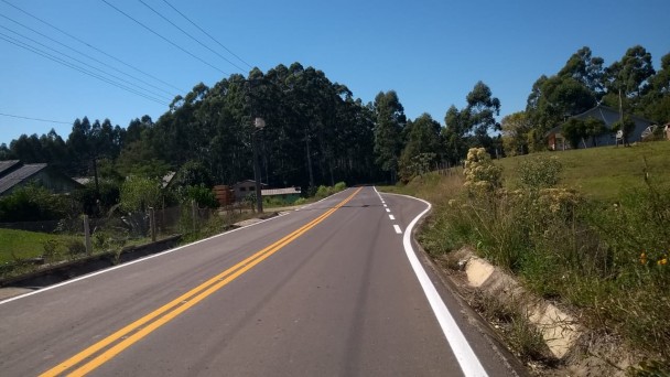 A foto mostra uma rodovia pavimentada com o asfalto sem imperfeições. No chão, ele está com a sinalização intacta. Ao redor, vegetação.