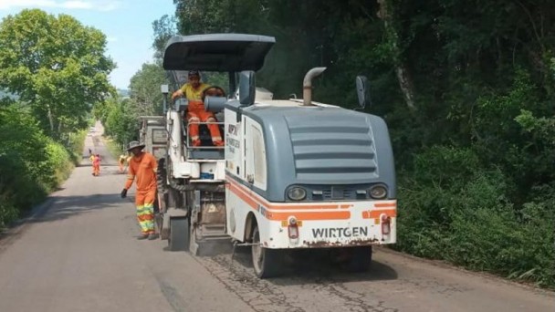 Na foto aparece uma máquina tratando o pavimento, com dois trabalhadores próximos a ela vestindo uma roupa laranja. Dos dois lados da imagem está a grande presença da vegetação. 