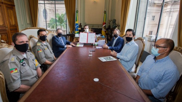 A foto se passa no Palácio Piratini. Ao fundo está o governador Eduardo Leite de máscara e tanto na parte direita quanto na esquerda, estão autoridades também utilizando máscaras de proteção. 