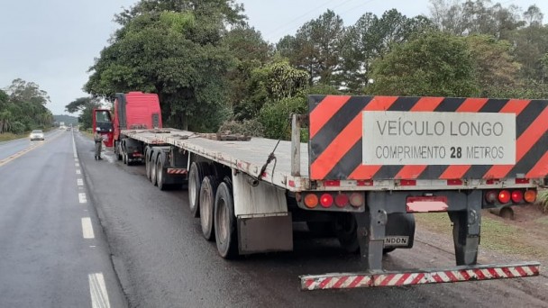 Na imagem está um caminhão longo de 28 metros. Ele é vermelho  e laranja e foi encostado na rodovia para ser fiscalizado. 
