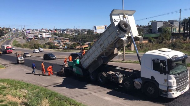 A imagem mostra um caminhão despejando o asfalto na pista, enquanto homens o espalham para que em seguida seja compactado