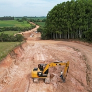 A foto mostra uma retroescavadeira em obras na rodovia de acesso ao município de Amaral Ferrador. A vista é panorâmica, captada por um drone.