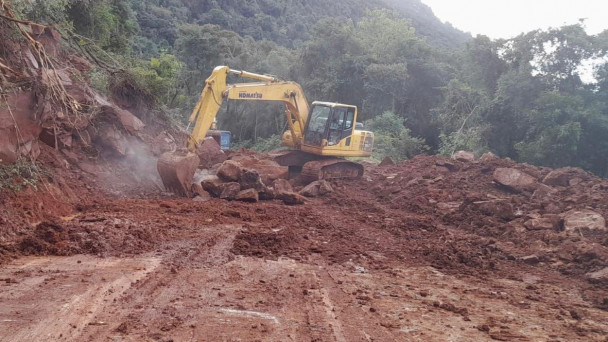 A foto mostra uma retroescavadeira retirando terras e rochas de um trecho interditado por deslizamento de encosta numa rodovia estadual gaúcha.