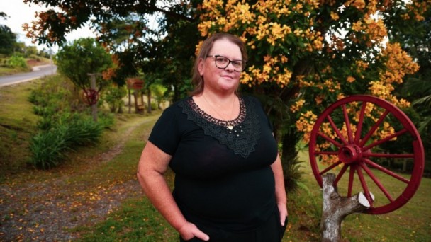 Imagem de Roselaine que mora junto à rodovia. Ela usa uma blusa preta e óculos com armação preta. Ela está em frente de uma árvore com flores amarelas e uma roda de carroça.